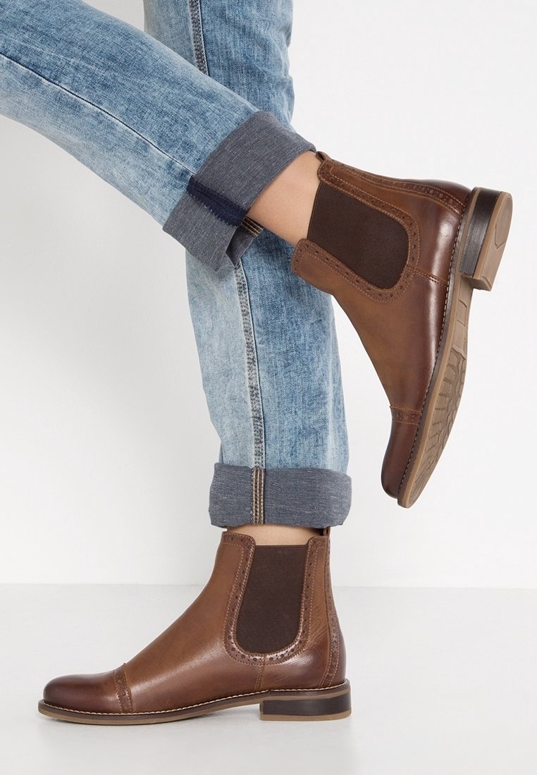 Bottes Chelsea en cuir marron avec panneaux latéraux élastiques portées avec un jean bleu clair retroussé sur un fond blanc.