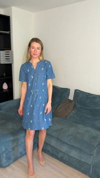 Γυναίκα με μπλε φόρεμα με κοντά μανίκια στέκεται ξυπόλυτη κοντά σε μία πετρόλ καναπέ με μαξιλάρια σε ένα σαλόνι με ανοιχτούς τοίχους.