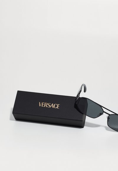 Mustat aurinkolasit tummilla linsseillä lepäävät mustalla teksturoidulla kotelolla. Kotelossa on kultakirjoitus brändin nimellä "VERSACE."