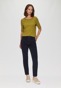 Olijfgroene geribbelde top met korte mouwen, gecombineerd met navy corduroy broek en beige sneakers met textuurdetails op een effen achtergrond.