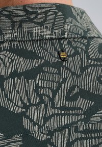 Groene textuurstof met een wit abstract patroon, met een klein zwart en geel "PME LEGEND" label nabij de kraagnaad.