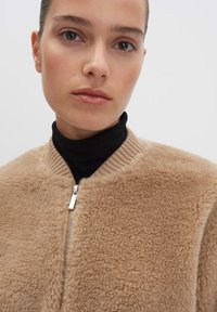 Giacca in pelliccia sintetica beige con finitura testurizzata, colletto a coste e dettagli in zip argentata. Indossata sopra un dolcevita nero.