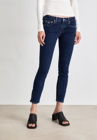 Vaqueros de mezclilla azul oscuro con corte slim, cintura de altura media, bolsillos frontales y detalle de cremallera en el dobladillo. Combinados con mules negros de punta abierta.