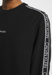 Felpa nera in cotone con scollatura a girocollo, caratterizzata da logo bianco "CALVIN KLEIN" lungo la cucitura della spalla e lungo le maniche. Texture morbida.