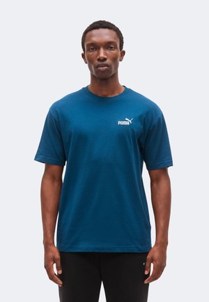 Uomo che indossa una maglietta blu Puma e pantaloni neri, in piedi rivolto verso davanti con un'espressione neutra su uno sfondo semplice.