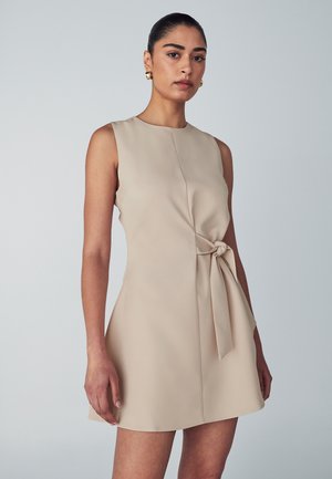 Robe sans manches beige avec un corsage ajusté, détail de taille noué et ourlet évasé. Texture lisse avec un design minimal, longueur au genou.