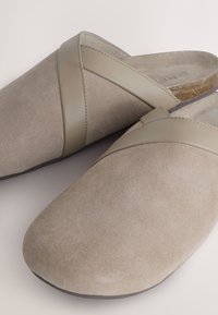 Chaussons beige sans fermeture avec devant en daim souple et lanières en cuir lisses croisées sur le dessus, semelle intérieure rembourrée et semelle en caoutchouc.