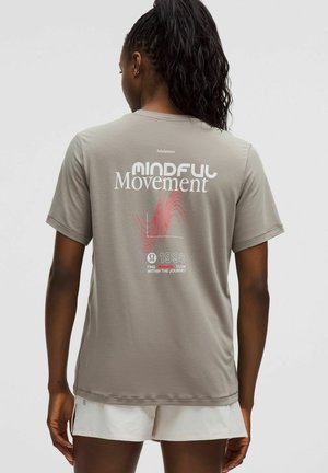 Frau mit dunklem, geflochtenem Haar trägt ein hellbraunes "Mindful Movement"-T-Shirt und weiße Shorts, steht mit dem Rücken zur Kamera.