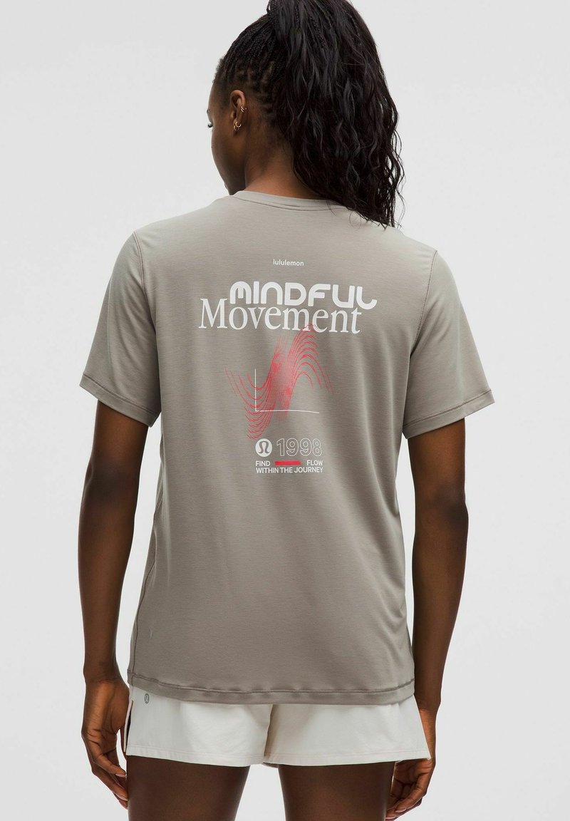 Tamsių pintų plaukų moteris vilki šviesiai rudos spalvos "Mindful Movement" marškinėlius ir baltas šortus, stovinti su nugara į kamerą.
