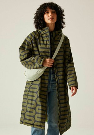 Kobieta o kręconych włosach ubrana w długi zielono-czarny płaszcz w wzory, niebieskie dżinsy oraz jasną torebkę na ramię, stojąca na tle jednolitego tła.