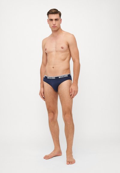Slip Moschino blu navy con cintura a contrasto bianca con logo, texture liscia e design aderente, mostrati su un modello maschile.