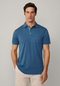 Mans polo skjorta i blått, med en tvåknapplåda, korta ärmar och en bröstficka. Mjuk tyg med en slät textur.