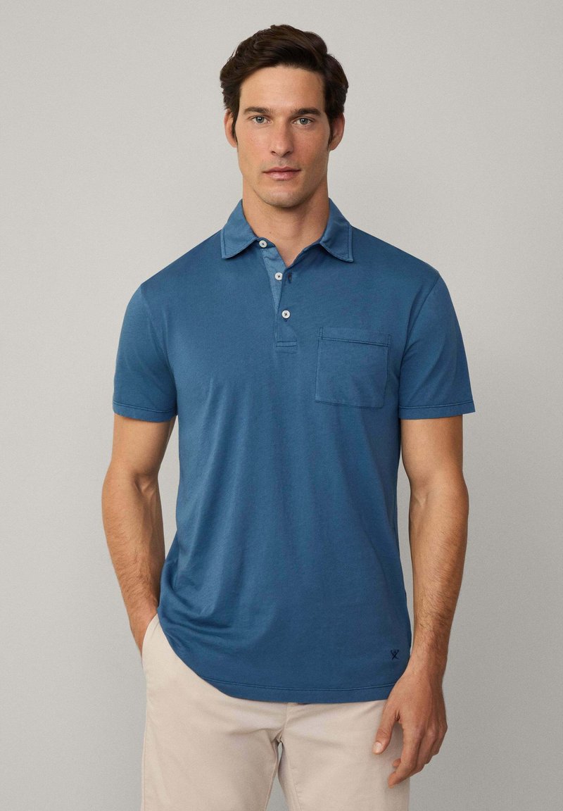 Mans polo skjorta i blått, med en tvåknapplåda, korta ärmar och en bröstficka. Mjuk tyg med en slät textur.