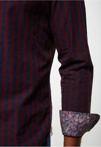 Camisa de manga larga con rayas verticales en rojo y azul marino. Los puños presentan un patrón floral colorido. Tejido suave y texturizado.