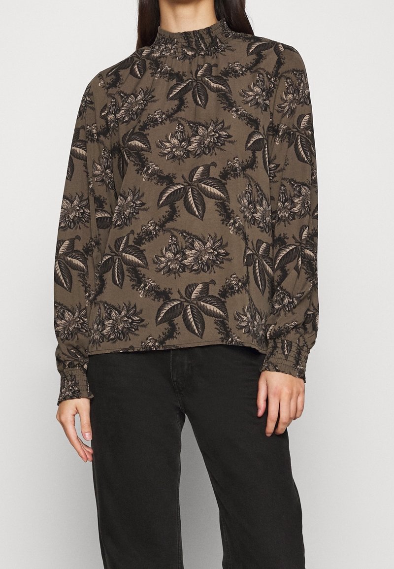 Frau trägt eine olivgrüne Bluse mit langen Ärmeln, schwarzem Blumendruck und gerafften Manschetten, kombiniert mit schwarzen Hosen.