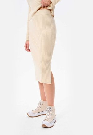 Rippierte, knielange cremefarbene Rock mit einem taillierten Taillenbund, kombiniert mit beigen High-Top-Sneakern mit weißen Akzenten und strukturierten Sohlen.