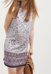 Bílé šaty bez rukávů s květinovým vzorem a námořnickými, červenými a růžovými detaily, s kulatým výstřihem a strukturovaným lemem.