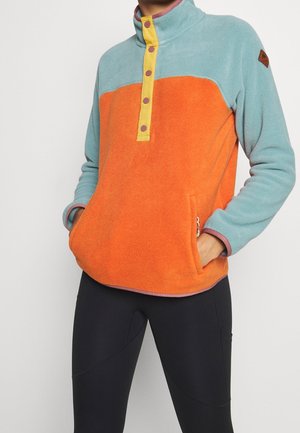 Osoba ubrana w polarową bluzę z blokowym wzorem kolorystycznym: jasnoniebieski górny fragment, pomarańczowe ciało, żółty pasek z guzikami oraz czarne legginsy, z rękami w przednich kieszeniach.