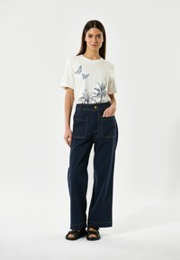 Hvid t-shirt med blå sommerfugl og palmetræ print, parret med mørkeblå brede bukser med gule knapper og synlige sømme.