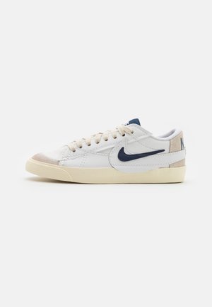 Baskets basses blanches avec semelle beige, logo Nike swoosh bleu marine et accents en daim beige au bout et au talon, vues de côté.