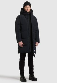 Czarna kurtka puchowa z zapięciem na zamek, kapturem i dwoma bocznymi kieszeniami, w połączeniu z czarnymi spodniami i masywnymi czarnymi butami.