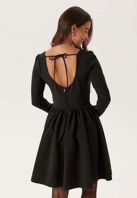 Zwarte lange mouwen jurk met een getailleerd bovenlijf, een uitlopende rok, een open rug en een strikdetail. Voorzien van een verborgen rits aan de achterkant.