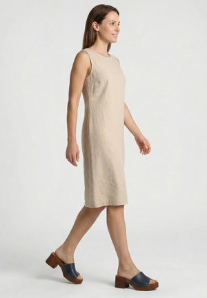 Kvinde går, iført ærmeløs beige knælængde kjole og marineblå åben tå blok-hælede træsko, på ensartet lys baggrund.