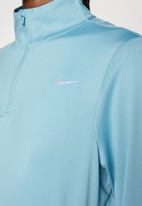 Lichtblauw sportieve quarter-zip top met een zachte textuur. Heeft een zilveren Nike-logo op de linkerkant van de borst en een hoge kraag. 