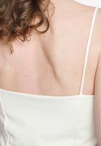 Gros plan du dos supérieur et de l'épaule d'une femme avec des cheveux bruns bouclés, portant un haut sans manches blanc avec de fines bretelles.