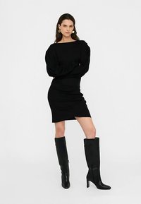 Sort figursyet kjole med puffede lange ærmer og firkantet udskæring, kombineret med høje sorte læderstøvler. Glat stof og knælængde design.