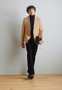 Tan švarkas virš juodos golfo megztinio, juodos kelnės ir rudos bateliai. Modelis laiko juodą rankinę, eidamas prieš paprastą foną.