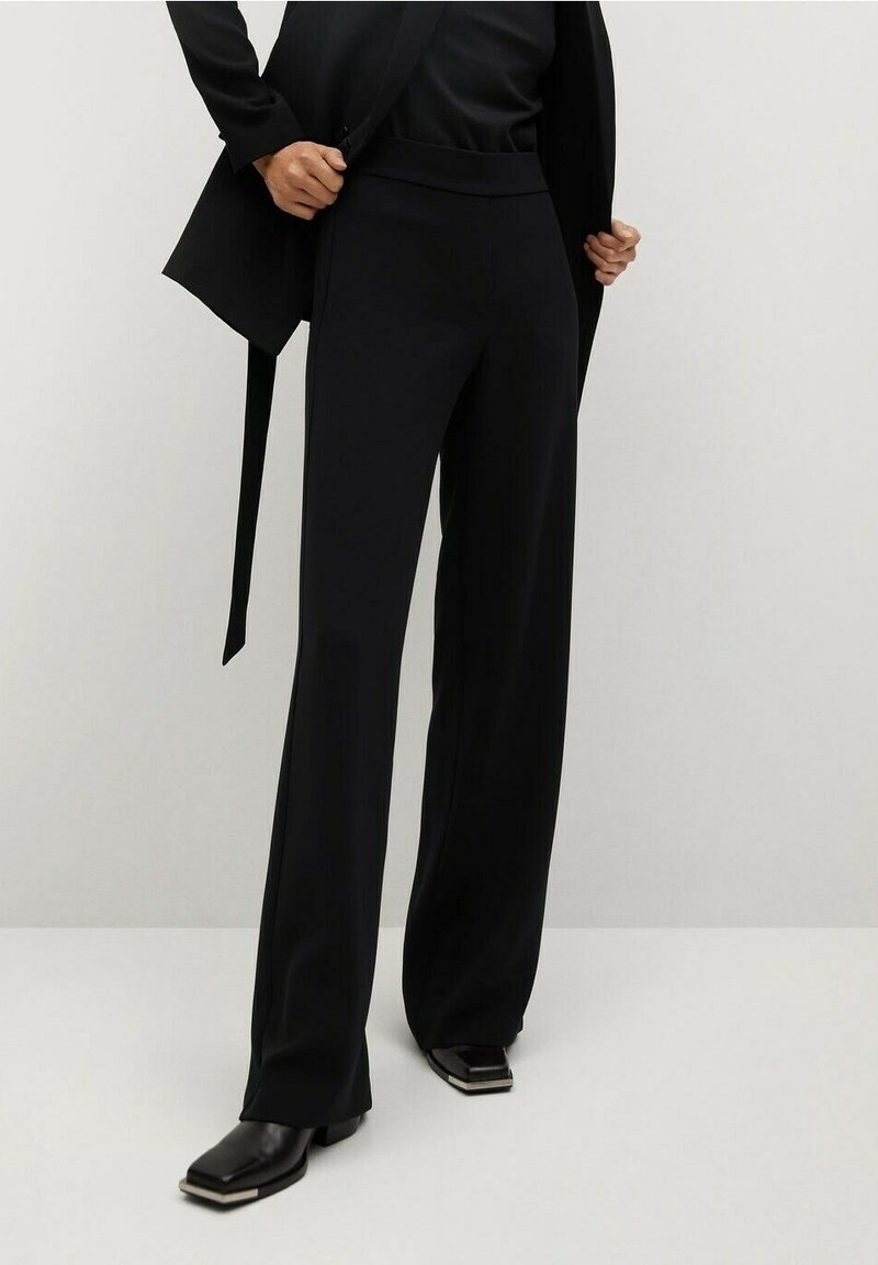 Pantalon large noir en tissu lisse avec une taille élastique, associé à un blazer noir bien taillé. Des chaussures brillantes notables créent un contraste.