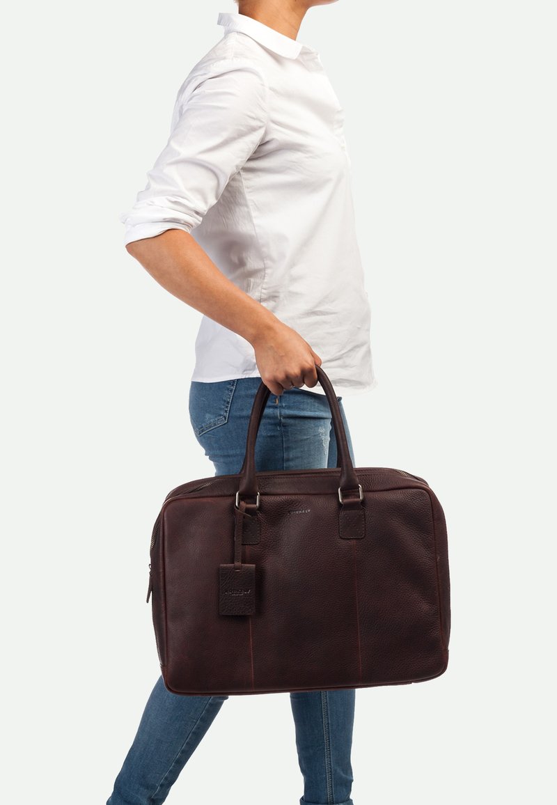 Borsa per laptop in pelle marrone con due manici robusti, chiusura con zip e etichetta per bagagli. Finitura testurizzata e forma rettangolare slanciata.