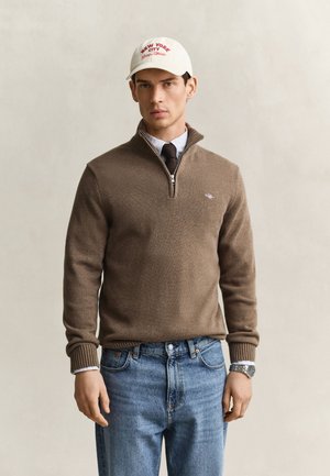 Homme portant un pull beige à demi-fermeture éclair, un jean bleu, une chemise blanche, une cravate noire, une montre argentée et une casquette blanche avec le texte rouge New York City.