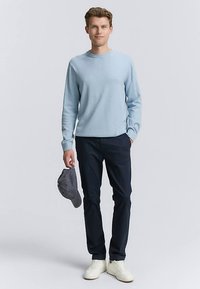 Svetlo moder pulover, temno modre hlače, siva kapa v roki in beli športni copati. Pulover ima dolge rokave in okrogel izrez.