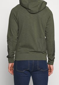 Olivově zelená mikina s lehkou tkaninou, dlouhými rukávy a kapsou typu klokanka. Má žebrované manžety a kapuci se stahovací šňůrkou.