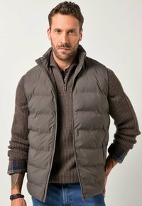 Chaleco marrón acolchado con un cuello alto sobre un suéter marrón de canalé, con un escote con cremallera y detalles de cuadros en los puños.