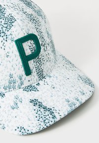Pet met een witte stof met een bloemenpatroon in blauw en groen. Heeft een groene geborduurde "P" aan de voorkant. Gebogen klep constructie.