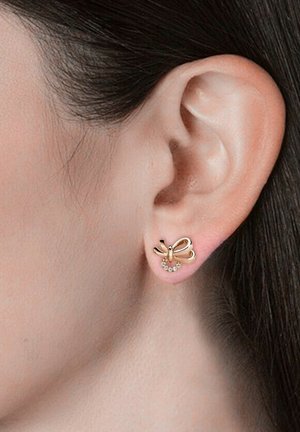 Orecchini color oro a forma di fiocco, con un piccolo design curvo e accenti di strass trasparenti, su uno sfondo di tonalità della pelle neutra.