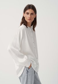 Camicia bianca con bottoni e maniche lunghe, in tessuto testurizzato e colletto rotondo. Abbinata a jeans in denim blu chiaro. Presenta polsini e bottoni.