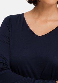 Donna che indossa una maglietta a maniche lunghe blu scuro con scollo a V, braccia incrociate, con i capelli castani corti visibili sul bordo dell'inquadratura.
