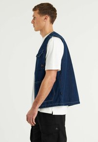 Denim jopica v temno modri barvi s sproščenim krojem, ki ima velike sprednje žepke in vidno šivane detajle. Nosi se preko bele majice.