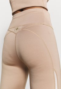 Ingrandimento di una persona che indossa leggings beige a vita alta con cuciture e un piccolo logo dorato sulla fascia posteriore.