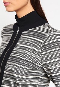 Veste à motifs noir et blanc avec un col montant, une fermeture zippée, et un tissu texturé à motifs géométriques.