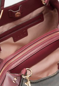 Interno di una borsa in pelle bordeaux, con tasca con cerniera e dettagli in oro, tasca aperta e fodera beige. Logo del marchio visibile.