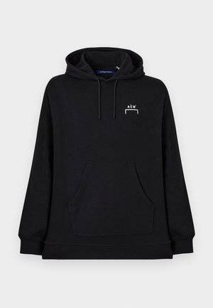 Joven de pie, vistiendo una sudadera negra con capucha y logo, pantalones negros de pierna ancha y zapatillas negras, mirando hacia un lado.