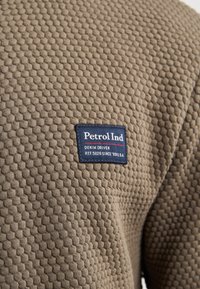 Texturerad brun tyg med ett honungskakemönster. En blå etikett med texten "Petrol Ind DENIM DRIVEN REF. 5026 SINCE '89 U.S.A."