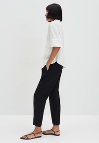 Witte textuurshirt met opgerichte mouwen, gecombineerd met zwarte taps toelopende broek en platte sandaaltjes met bandjes, gezien in profiel tegen een lichte achtergrond.