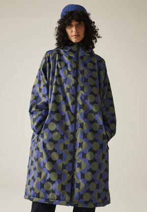 Kobieta o kręconych włosach, ubrana w wzorzysty niebiesko-zielony długi płaszcz i niebieską dzianinową czapkę, stoi z rękami w kieszeniach płaszcza.