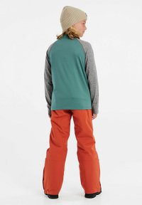 Maglietta a maniche lunghe verde e grigia con scollo tondo, abbinata a pantaloni arancioni vivaci. Il materiale sembra leggero e flessibile, con una vestibilità comoda.